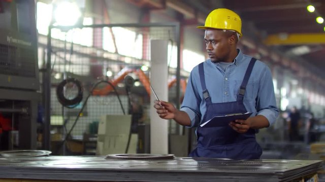 Medium pan shot of black mechanic dressed in blue overalls and safety helmet filling out paperwork, counting batch of finished gear wheels, with metalwork machines and power tools in background