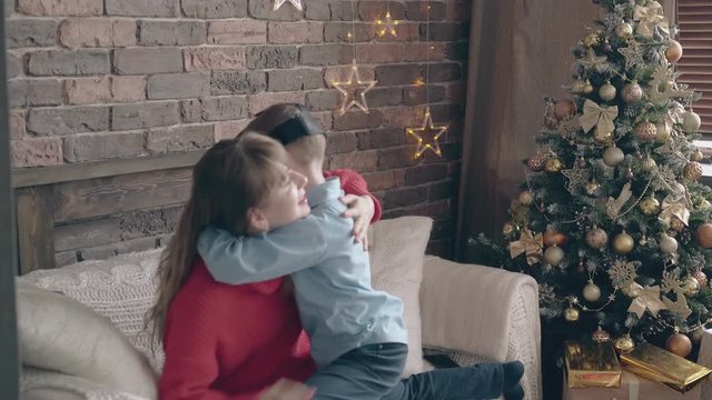son with VR headset hugs tight pretty mom on white settee near new year tree against brick wall with LED stars