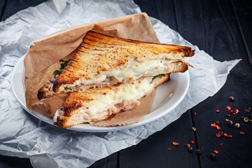 Close up on grilled sandwich with salmon and melted cheese and lettuce. Snack. Fast food for lunch. Sandwich served on white plate on dark wooden background