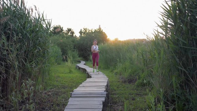 running in nature, active sports woman with beautiful body in fitness clothes engaged jogging on wooden bridge outdoors among green reeds at sunset