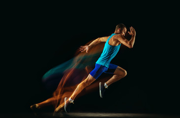 Professional male runner training isolated on black studio background in mixed light. Man in sportsuit practicing in run or jogging. Healthy lifestyle, sport, workout, motion and action concept. - obrazy, fototapety, plakaty
