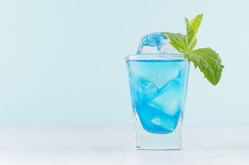 Bright summer fresh blue fruit cocktail with blue curacao liquor, ice cubes,  green mint in pastel mint color interior on white wood board.