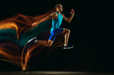 Professional male runner training isolated on black studio background in mixed light. Man in sportsuit practicing in run or jogging. Healthy lifestyle, sport, workout, motion and action concept.