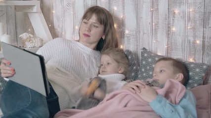 kids watch cartoons on gray tablet lying on bed among pillows with mother at wooden wall and LED fairylights in room