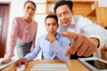 Mature Asian businessman pointing at computer screen of coworker, focus on index finger