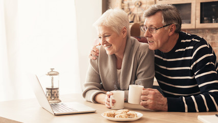 Elderly retired couple reading their social media news on laptop in kitchen