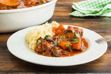 Coq au Vin, traditional French recipe of chicken braised in red wine with carrot and mushrooms. Served with mashed potatoes. White casserole and plate on wooden table