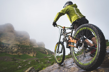 Rear view wide angle of a man on a mountain bike stands on a rocky terrain and looks at a rock. The...