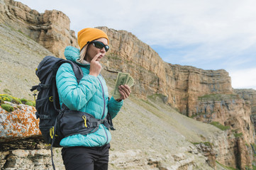 A girl traveler in sunglasses and with a backpack on the background of high mountain rocks thoughtfully holds one hundred dollar bills. Finance concept for tourism and travel vouchers