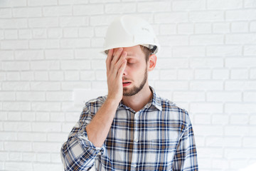 A blue collar worker wearing a hardhat is covering his face with his hand