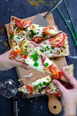 Flatbread healthy pizza slices with chicken, tomatoes, pepper, mozzarella on a wooden cutting board. piece of pizza in a hand. proper nutrition on dark background. top view
