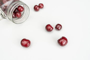 Red juicy cherries scattered from out the glass jar on a white table from the upper-left corner