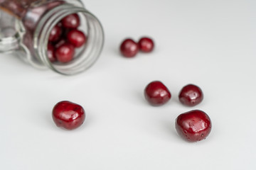 Red juicy cherries scattered from out the glass jar on a white table 