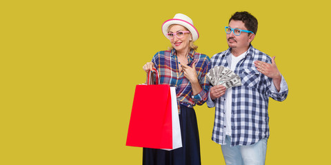 Happy family after shopping, adult man and woman in casual checkered shirt standing together,wife holding paper bag with toothy smile,husband fan of money like big sponsor.Indoor, isolated,studio shot