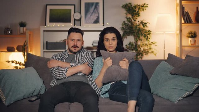 Husband and wife watching scary movie on TV looking at camera with scared faces holding pillows sitting on sofa in apartment. Youth and emotions concept.
