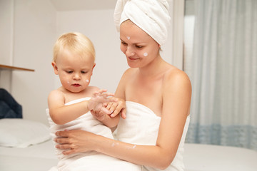 Mother and infant baby in white towels after bathing apply sunscreen or after sun lotion or cream. Children skin care