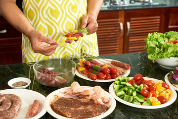 Close-up of young man in apron holding kebab with meat and fresh vegetables and cooking the other ones for barbecue