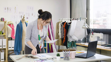 Young people small business. asian woman at work as fashion designer and tailor looking at sketches of new collection in atelier on desk. dressmaker lady in eyeglasses concentrated in studio workshop