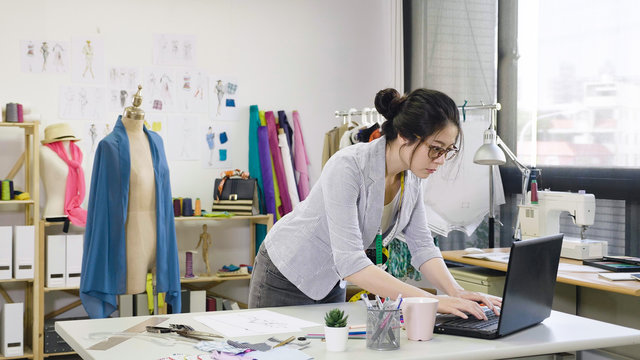 Responding on business e mail. Beautiful young Asian chinese woman working using computer and smiling while standing in workshop. fashion designer discussing with customer customize dress making.