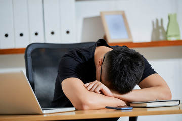 Young businessman in casual black T-shirt sleeping at his workplace with laptop and notepad during work day at office