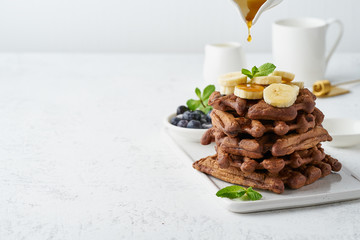 Chocolate banana waffles with maple syrup on white table, copy space, side view. Sweet brunch,...