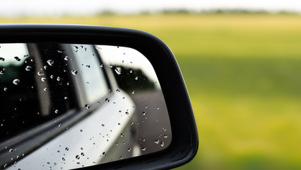 raindrops on side rear-view mirror on a car in a raining day. drops of rain on car window. sunset, sun light, green field over rainy wet road. driving in bad weather.