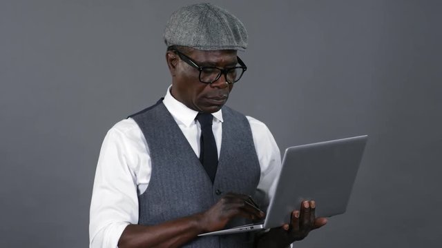Middle aged african american businessman in tweed cap and elegant vest and shirt standing in studio against grey background and using laptop