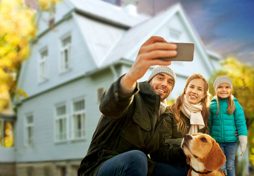 family, pets and people concept - happy mother, father and little daughter with beagle dog taking selfie by smartphone outdoors over house in autumn background