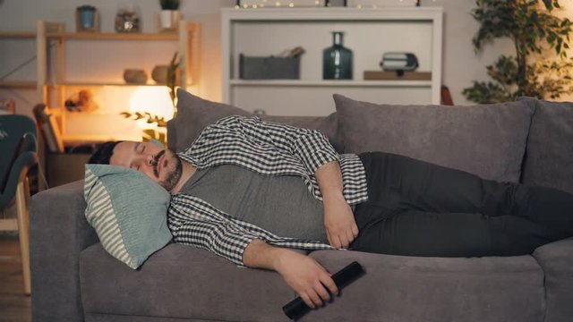 Young man is sleeping on couch at home holding remote control for television resting late at night in cozy illuminated room. People and lifestyle concept.
