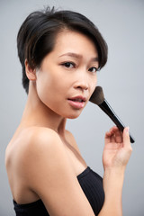 Portrait of Asian beautiful woman looking at camera while applying powder on her face with make-up brush isolated on grey background