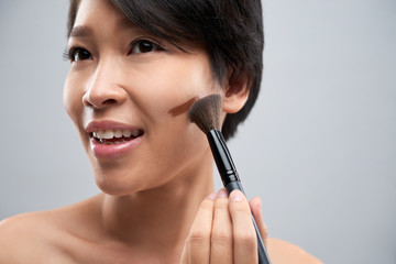 Close-up of Asian smiling woman applying tone cream on her face with make-up brush
