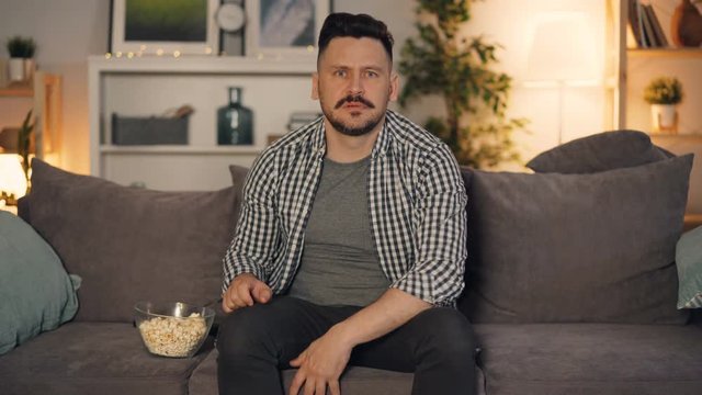 Guy is watching interesting TV program at home eating popcorn gesturing and talking commenting content on screen. Modern entertainment and people concept.