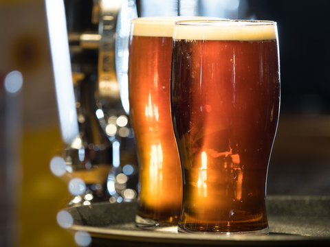 Two pints of dark coloured real ale stand on a tray in pint glasses with frothy heads back lit on a bar top. - Image