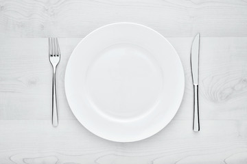 Empty white round ceramic plate, fork and knife on white wooden table with copy space