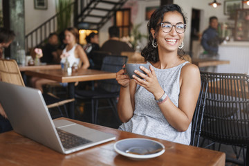 Girl taking moment relax. Happy woman curly short haircut resting working laptop sit cafe indoors lean rattan chair contemplate summer view outside window smiling hold cup cappuccino take break