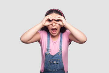 Portrait of amazed young girl in pink t-shirt and blue overalls standing with binoculars gesture hands on eyes and looking with shocked face. indoor studio shot, isolated on light gray background.