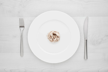 Small portion of tagliatelle pasta on round white plate with fork and knife on a white wooden table. Concept of dieting and healthy eating.