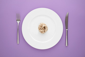 Small portion of tagliatelle pasta on round white plate with fork and knife on light purple tablecloth. Concept of dieting and healthy eating.