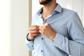 Man buttons sleeves on blue linen dress shirt in bedroom at slight angle.