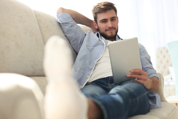 Handsome man is using a digital tablet and smiling while resting on couch at home.