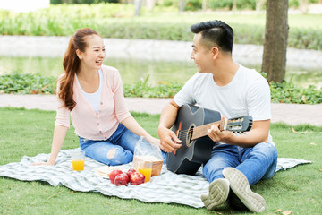 Asian young man playing on guitar to his girlfriend while she sitting and laughing during summer picnic in the park