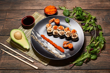 Fototapeta na wymiar Original Japanese rolls in restaurant serving with fish slices. Avocado, soy sauce, caviar, salad leaves, chopsticks around black plate with sushi. Bamboo napkin on wooden dark background. Top view.