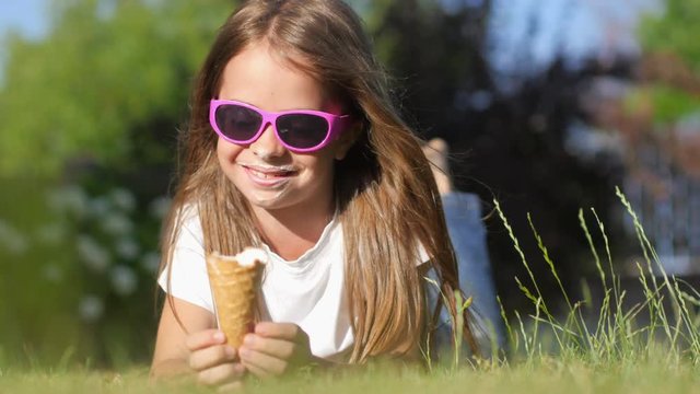 Beautiful little girl in sunglasses with a tasty ice cream lies on the lawn in the park and enjoys. The child eats ice cream with a dirty face. 4K