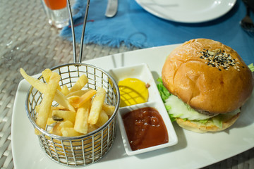 French fries in stainless steel basket with sauce and hamburger on white dish
