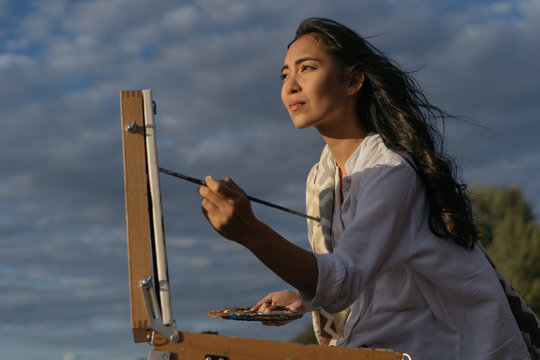 Asian woman artist painting picture on canvas outdoor. Portrait of cheerful Korean painter holding paintbrush and palette, admire beautiful landscapes and amazing sunset in park. Art education