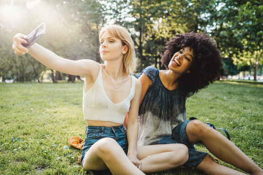 Two young women takes a selfie with the smart phone - Millennial relaxing at the park at sunset on a summer day