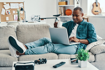 Happy to rest. Handsome young African man using laptop and smiling while sitting indoors
