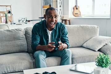 So much fun! Handsome young African man playing video game and smiling while sitting indoors
