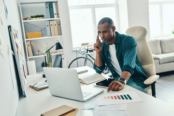 Fototapeta premium Concentrated at work. Thoughtful young African man in shirt talking on the smart phone while sitting in the office