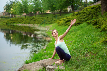 Obraz na płótnie Canvas Slim young brunette yogi performs no complicated yoga exercises on the green grass in summer against the background of nature